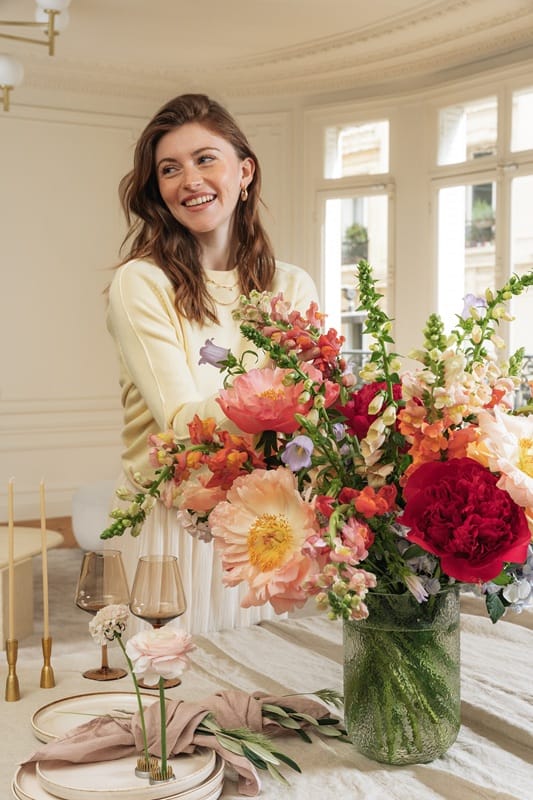 Femme souriante derrière un gros bouquet de fleurs posé sur une table dans un appartement